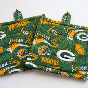 May include: Two square fabric pot holders featuring a repeating Green Bay Packers pattern in green, yellow, and white. The design includes football helmets, footballs, pennants, and the team name text. Each pot holder has a small fabric hanging loop.