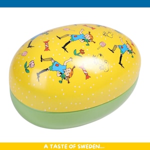 Può includere: Scatola a forma di uovo gialla con base verde, decorata con illustrazioni di una bambina con un vestito blu, fiori e caramelle. La scatola ha un motivo a pois e il testo "A TASTE OF SWEDEN...." in basso.