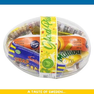 Puede incluir: Un recipiente de plástico transparente lleno de varios caramelos y golosinas, incluyendo envoltorios naranjas y amarillos. Una banda amarilla con el texto "Gled Pus" envuelve el recipiente. El texto "A Taste of Sweden" está en la parte inferior.