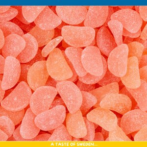Pode incluir: Close-up de uma pilha de gomas laranja e rosa, em forma de fatias de laranja. As gomas são revestidas com açúcar e têm uma aparência translúcida. O texto "A TASTE OF SWEDEN...." está na parte inferior.