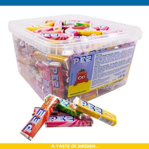 May include: A clear plastic container filled with colorful PEZ candy dispensers and refills. Several dispensers are scattered in front, showcasing various fruit flavors. The container has a label with text and a cartoon strawberry.