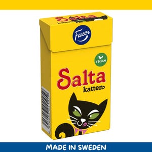 Könnte beinhalten: Eine gelbe Schachtel mit veganen Fazer Salta Katten Süßigkeiten. Die Schachtel zeigt eine schwarze Katze mit grünen Augen und einer rosa Schleife. Der Text "Salta katten" ist rot, und das Fazer-Logo befindet sich oben.