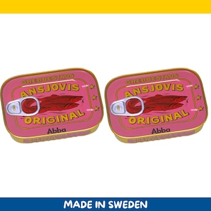 Puede incluir: Dos latas rectangulares rosas de Grebbestads Ansjovis Original, con una anilla y un gráfico de peces rojos. Las latas tienen ribetes dorados y la marca Abba está impresa en la parte inferior. El texto "MADE IN SWEDEN" está en la parte inferior.