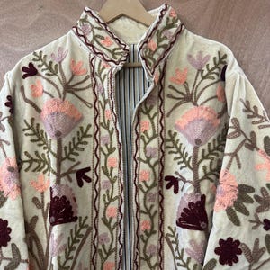 May include: Cream-colored embroidered jacket with a high collar. The jacket features a floral design in shades of pink, burgundy, and green. The lining is striped. The jacket is hanging on a wooden hanger.