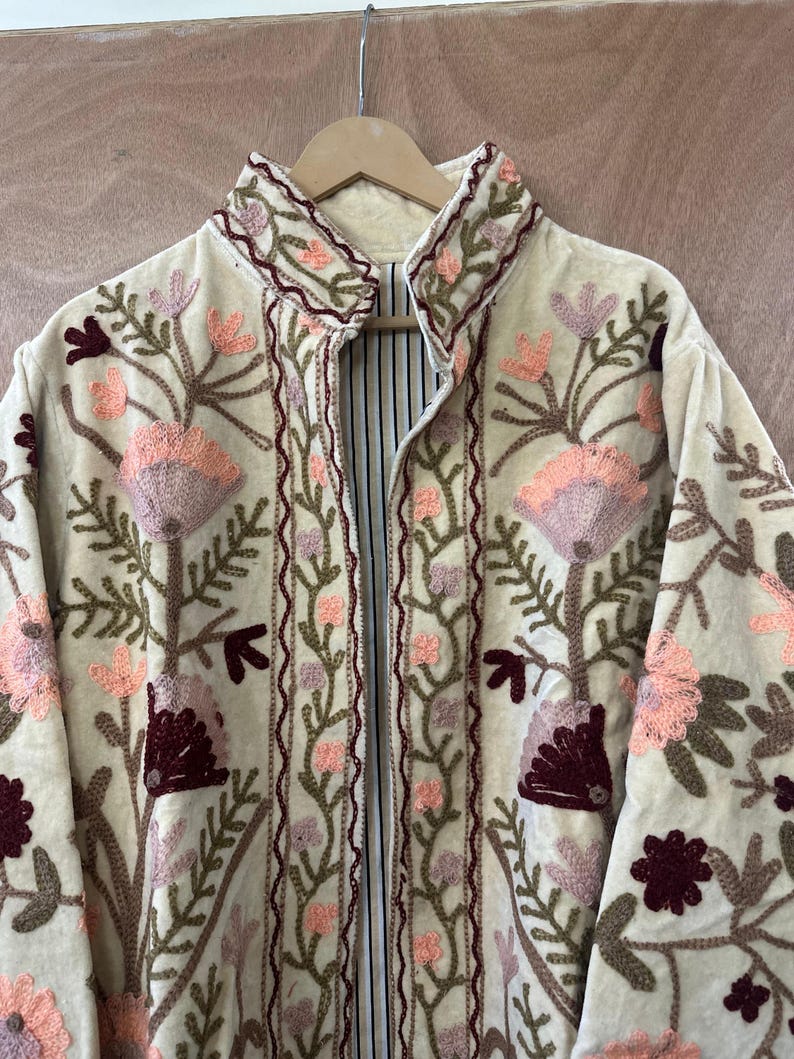 May include: Cream-colored embroidered jacket with a high collar. The jacket features a floral design in shades of pink, burgundy, and green. The lining is striped. The jacket is hanging on a wooden hanger.