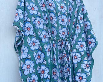 May include: A green floral print kaftan dress. The dress features a repeating pattern of white and blue flowers with orange centers. The kaftan has a V-neck and a drawstring waist. The dress is hanging on a wooden hanger.