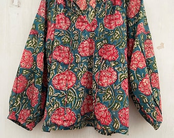 May include: A long-sleeved blouse with a floral pattern. The blouse features a teal background with large red flowers and green leaves. The top has a V-neck and is hanging on a wooden hanger.