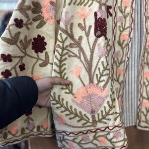 May include: Cream-colored embroidered jacket with floral and leaf patterns in shades of pink, brown, and green. The jacket features a striped panel and intricate embroidery along the edges. The design includes a variety of flowers and foliage.