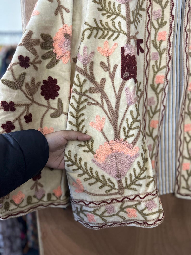 May include: Cream-colored embroidered jacket with floral and leaf patterns in shades of pink, brown, and green. The jacket features a striped panel and intricate embroidery along the edges. The design includes a variety of flowers and foliage.