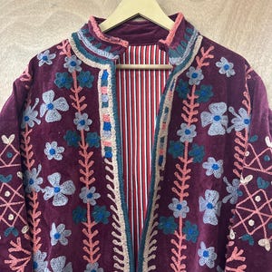 May include: A burgundy velvet jacket with floral embroidery. The jacket features a high collar and a striped red and white lining. The embroidery includes blue, gray, and pink flowers and decorative patterns. The jacket is hanging on a wooden hanger.