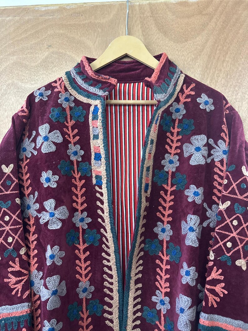 May include: A burgundy velvet jacket with floral embroidery. The jacket features a high collar and a striped red and white lining. The embroidery includes blue, gray, and pink flowers and decorative patterns. The jacket is hanging on a wooden hanger.