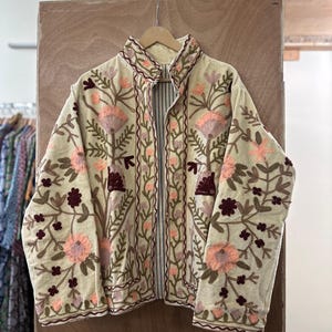 May include: Cream-colored jacket with intricate floral embroidery in shades of pink, brown, and green. The jacket features a high collar and a full-length zipper. The embroidery is detailed and covers the front, sleeves, and collar. The text "IS:1659" is visible.