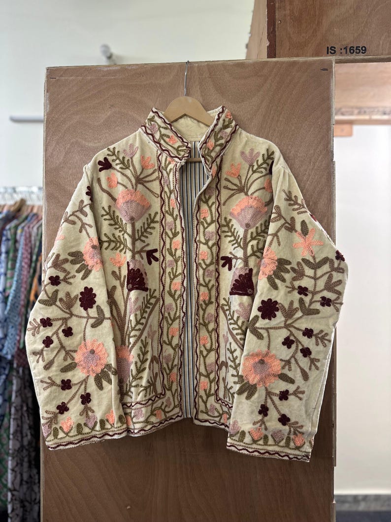 May include: Cream-colored jacket with intricate floral embroidery in shades of pink, brown, and green. The jacket features a high collar and a full-length zipper. The embroidery is detailed and covers the front, sleeves, and collar. The text "IS:1659" is visible.