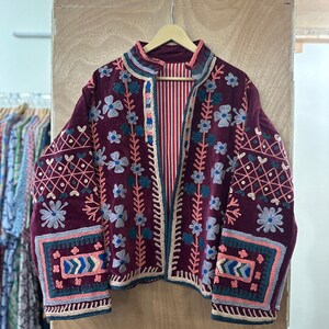 May include: A burgundy embroidered jacket with floral and geometric patterns. The jacket features a collarless design, long sleeves, and a striped red and white lining. The embroidery includes shades of blue, pink, and teal, creating a bohemian aesthetic.