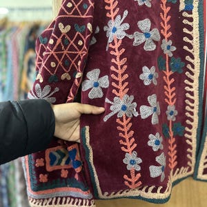 May include: Burgundy velvet jacket with floral embroidery in white, blue, and peach. The jacket features intricate patterns on the sleeves and edges, with a crochet trim. The design is a blend of textures and colors.