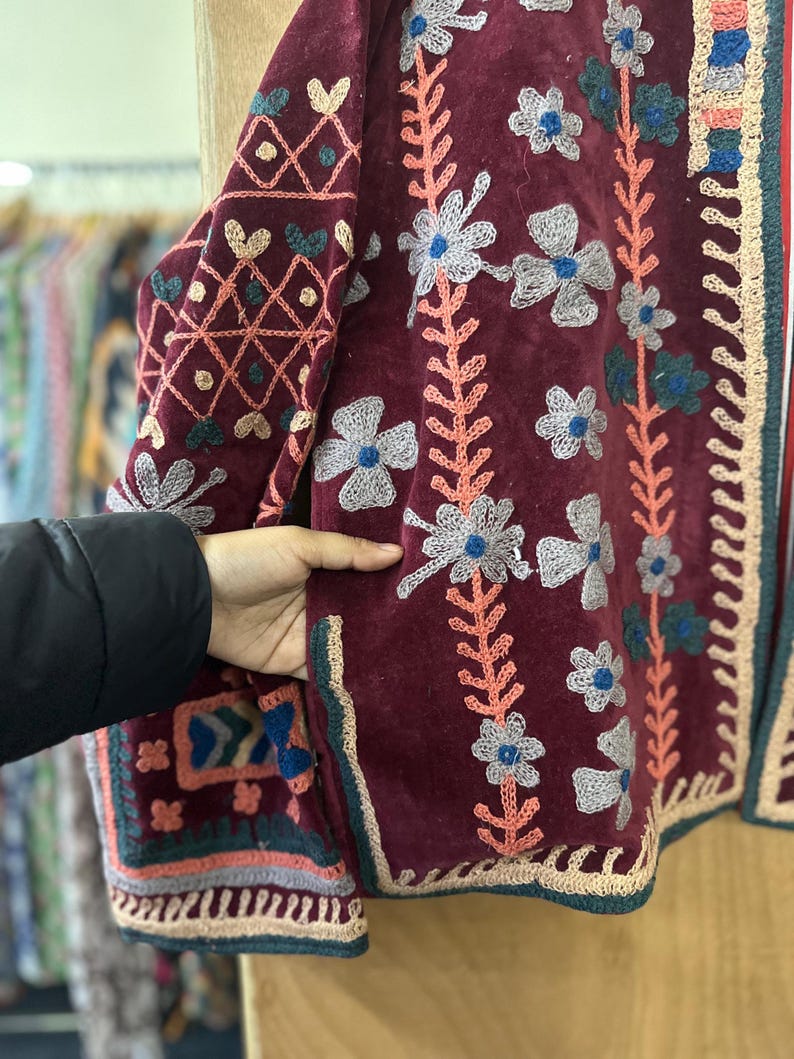 May include: Burgundy velvet jacket with floral embroidery in white, blue, and peach. The jacket features intricate patterns on the sleeves and edges, with a crochet trim. The design is a blend of textures and colors.