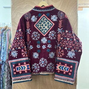 May include: Burgundy jacket with intricate floral and geometric embroidery in shades of pink, blue, and white. The jacket features a high collar and detailed patterns on the sleeves and back, creating a bohemian aesthetic.