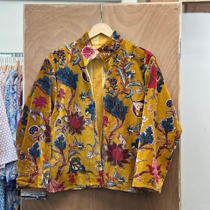 Puede incluir: Chaqueta de color amarillo mostaza con un vibrante estampado floral en rojo, azul y verde. La chaqueta, sin cierre, cuelga de una percha de madera. El diseño floral cubre toda la chaqueta.