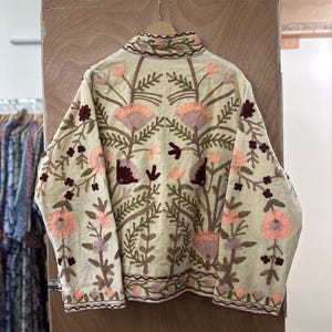 May include: Cream-colored jacket with intricate floral embroidery in shades of peach, burgundy, and green. The jacket features a high collar and detailed patterns across the front and sleeves. The embroidery is a prominent design element.