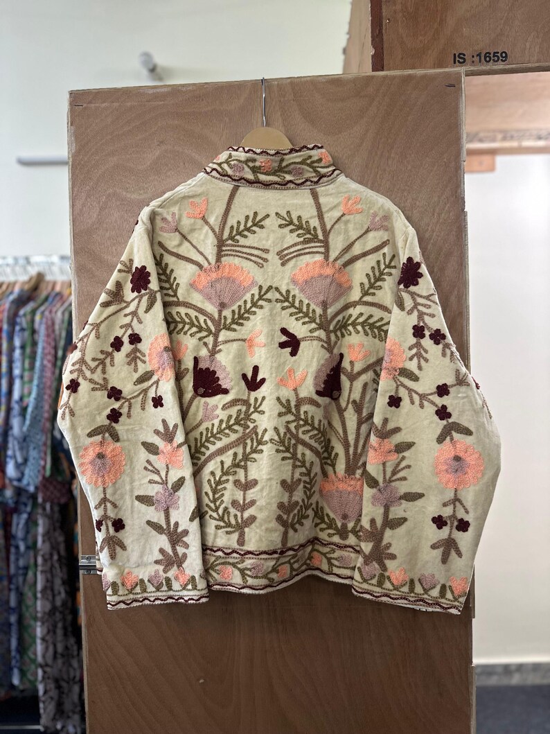 May include: Cream-colored jacket with intricate floral embroidery in shades of peach, burgundy, and green. The jacket features a high collar and detailed patterns across the front and sleeves. The embroidery is a prominent design element.