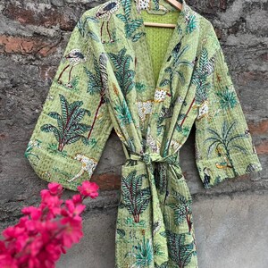 May include: A light green robe with a jungle pattern featuring zebras, leopards, and birds. The robe has long sleeves and a tie waist. It is hanging on a wooden hanger. The background is a textured brick wall.