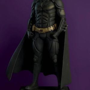 May include: A detailed Batman figurine standing on a black rectangular base with the text THE DARK KNIGHT embossed on the front. The figure wears a matte black armored suit with a gold utility belt, a long flowing cape, and a pointed cowl mask.