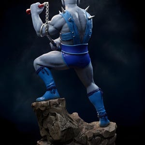 Puede incluir: Figura de acci&oacute;n de piel azul de un personaje musculoso, posando sobre una base rocosa. La figura lleva pantalones cortos azules, botas y un arn&eacute;s con p&uacute;as plateadas. Sostiene un arma de cadena con mangos rojos, sobre un fondo oscuro.
