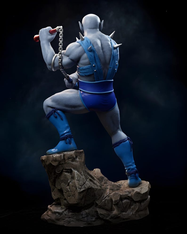 Puede incluir: Figura de acci&oacute;n de piel azul de un personaje musculoso, posando sobre una base rocosa. La figura lleva pantalones cortos azules, botas y un arn&eacute;s con p&uacute;as plateadas. Sostiene un arma de cadena con mangos rojos, sobre un fondo oscuro.