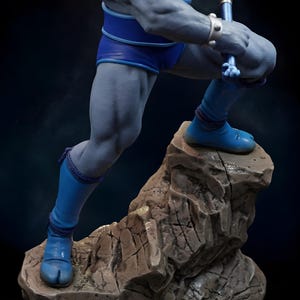 Puede incluir: Estatua detallada de una figura musculosa de piel gris, con pantalones cortos, botas y una parte superior azules. La figura posa sobre una base rocosa, sosteniendo un arma. La estatua tiene detalles intrincados y un fondo oscuro.