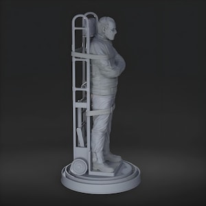 May include: A gray sculpture of a person in a straight jacket, secured to a wheeled frame. The figure is wearing a mask and is standing on a circular base. The frame has a ladder-like structure.