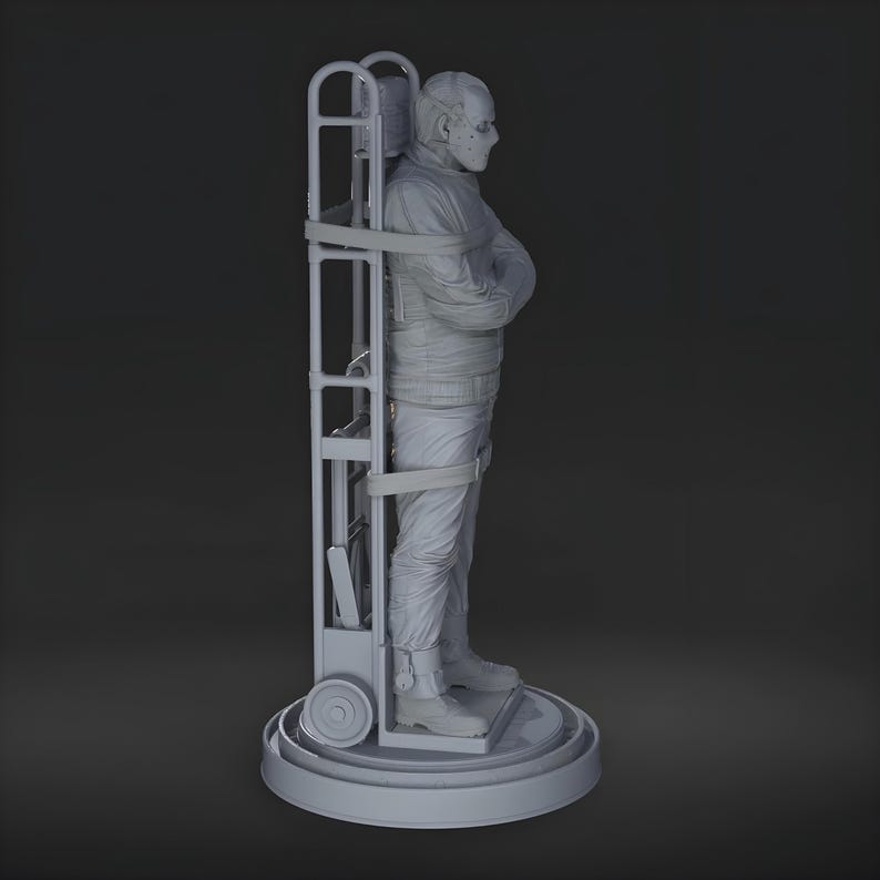 May include: A gray sculpture of a person in a straight jacket, secured to a wheeled frame. The figure is wearing a mask and is standing on a circular base. The frame has a ladder-like structure.