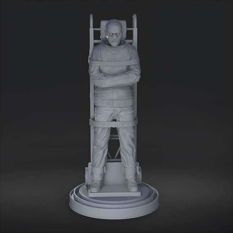 May include: A gray sculpture of a person strapped to a restraining chair. The figure is wearing a long-sleeved shirt and pants, with straps across the chest, waist, and legs. The chair has a circular base and a ladder-like back.