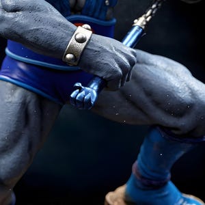 Puede incluir: Primer plano de una figura musculosa de piel gris, con pantalones cortos y botas azules, sosteniendo un arma azul. La figura lleva una pulsera plateada con tachuelas. La imagen est&aacute; sobre un fondo oscuro.