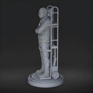 May include: A gray sculpture of a person in a straight jacket, secured to a wheeled cart. The figure stands on a circular base, arms crossed, with a neutral expression. The cart has a ladder-like frame.