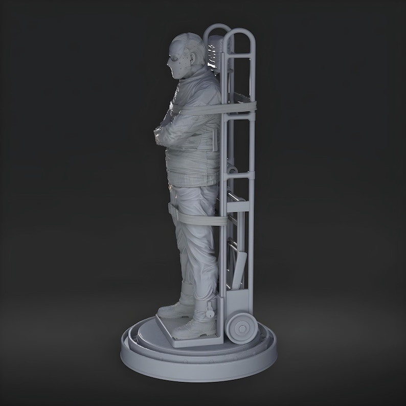 May include: A gray sculpture of a person in a straight jacket, secured to a wheeled cart. The figure stands on a circular base, arms crossed, with a neutral expression. The cart has a ladder-like frame.