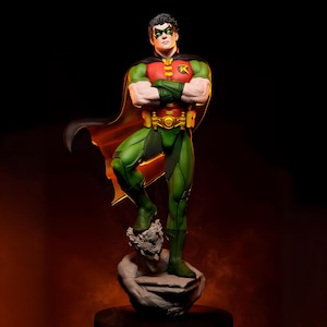 May include: A detailed statue of Robin, a DC Comics superhero, in a dynamic pose. The figure wears a green and red costume with a black cape. The statue is standing on a stone base with a sculpted face. The background is dark with an orange glow.