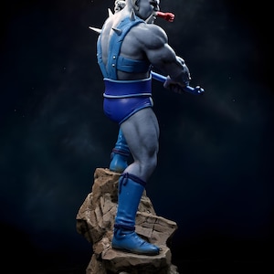 Puede incluir: Figura detallada de un personaje musculoso de piel gris en una pose din&aacute;mica. La figura lleva pantalones cortos azules, botas y un arn&eacute;s con p&uacute;as. Se encuentra sobre una base rocosa y sostiene un arma azul, con un objeto rojo en la boca.