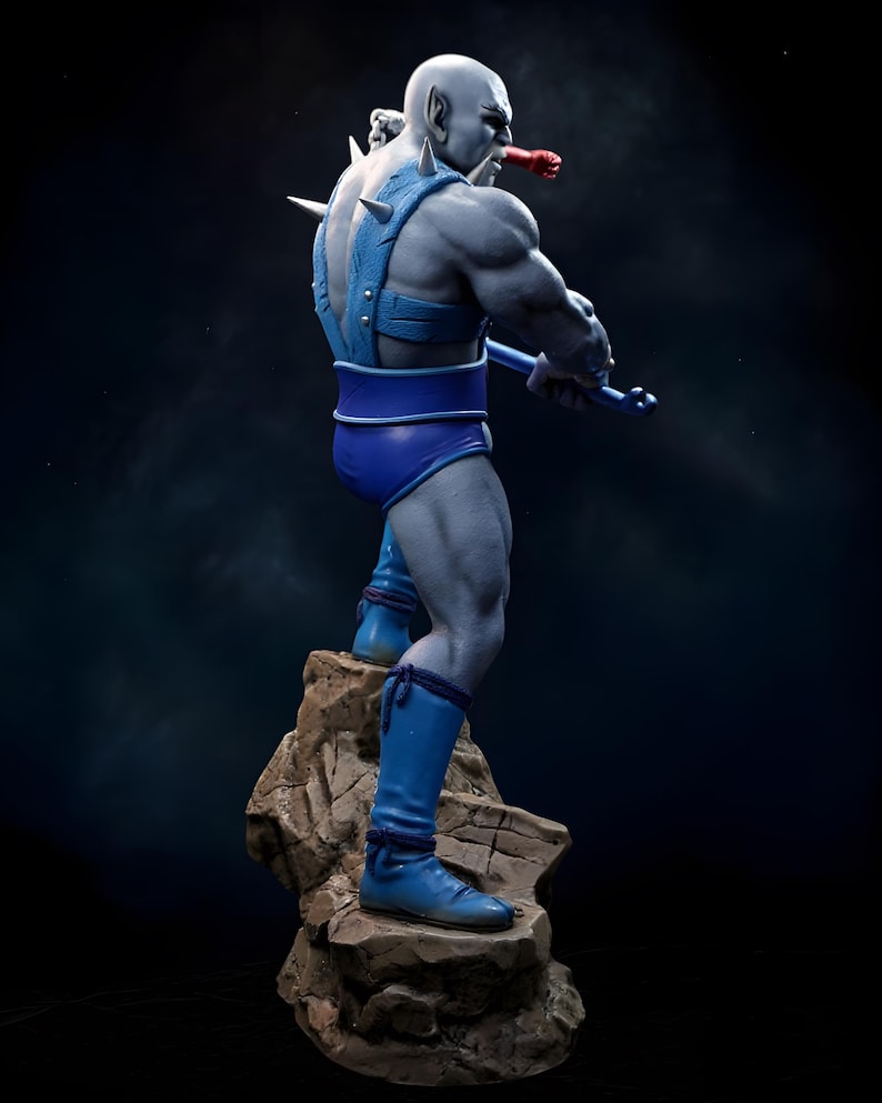 Puede incluir: Figura detallada de un personaje musculoso de piel gris en una pose din&aacute;mica. La figura lleva pantalones cortos azules, botas y un arn&eacute;s con p&uacute;as. Se encuentra sobre una base rocosa y sostiene un arma azul, con un objeto rojo en la boca.