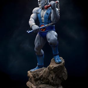 Puede incluir: Figura detallada de un personaje humanoide musculoso de piel gris, posando sobre una base rocosa. La figura viste un atuendo azul con un cintur&oacute;n con un emblema naranja. Sostiene un arma de cadena con detalles rojos y azules.