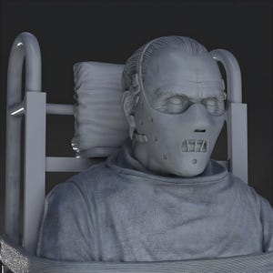 May include: A gray sculpture of a person wearing a face mask and a gray smock, seated in a chair with a headrest. The mask has multiple holes and a mouth guard. The sculpture has detailed facial features and is set against a dark background.
