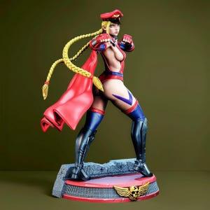 May include: A collectible figurine of a blonde character with long braided hair wearing a red military-style cap, a red bodysuit with blue accents, red gloves, and black knee-high boots. The figure stands on a circular base with a skull emblem.