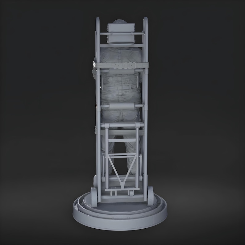 May include: A gray figure in a jumpsuit is secured within a gray hand truck. The hand truck has a rectangular frame with wheels and a platform base. The figure is facing away from the viewer, with the back of the jumpsuit visible.