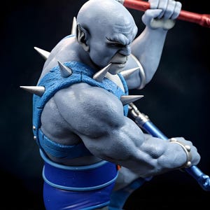 Puede incluir: Figura detallada de una figura humanoide musculosa de piel azul. La figura est&aacute; vestida con una armadura azul con p&uacute;as plateadas y sostiene un bast&oacute;n rojo y azul. El personaje tiene una expresi&oacute;n severa y est&aacute; en una postura de acci&oacute;n din&aacute;mica.
