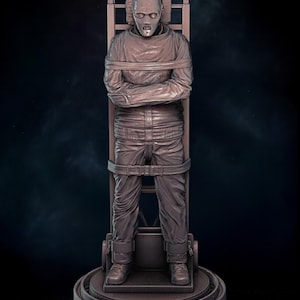 May include: A brown sculpture of a person strapped to a wooden chair. The person is wearing a long-sleeved shirt and pants, with their arms crossed. The sculpture has a detailed, realistic appearance and is set against a dark background.