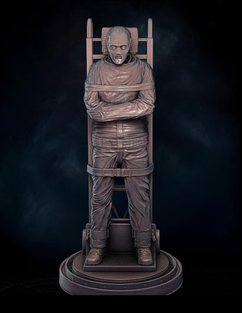 May include: A brown sculpture of a person strapped to a wooden chair. The person is wearing a long-sleeved shirt and pants, with their arms crossed. The sculpture has a detailed, realistic appearance and is set against a dark background.