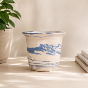May include: A ceramic planter featuring a cream-colored base with a swirling blue marbled pattern. The vessel has a wide, flared rim and a rounded body, showcasing a smooth finish with horizontal blue streaks concentrated near the base.
