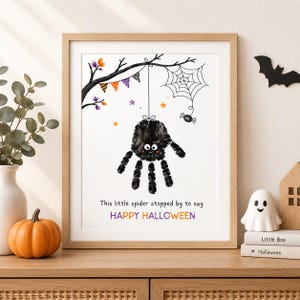 May include: A framed art print featuring a black handprint spider hanging from a tree branch with a web, colourful bunting, and small stars. The text reads: This little spider stopped by to say HAPPY HALLOWEEN in black and orange lettering.