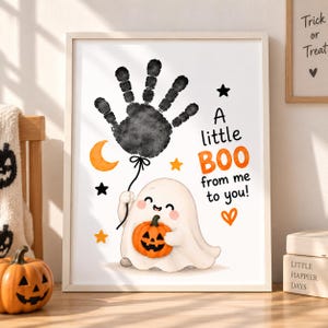 Può includere: Una stampa artistica incorniciata con un palloncino a forma di impronta di mano nera tenuto da un fantasma bianco che regge una piccola zucca arancione. Il testo recita A little BOO from me to you! con stelle arancioni e nere, una luna.