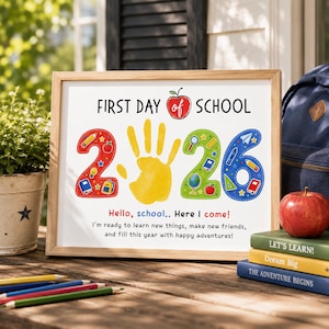 Pode incluir: Um cartaz emoldurado com o texto FIRST DAY OF SCHOOL e 2026, apresentando uma impressão de mão amarela no centro. Os números são decorados com material escolar. Abaixo, o texto diz Hello, school.. Here I come! e I'm ready to learn.
