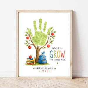 Può includere: Una stampa incorniciata con un albero a forma di impronta di mano, foglie verdi e mele rosse. Sotto l'albero ci sono libri, una matita gialla e uno zaino blu con un uccellino blu. Il testo recita: Watch me GROW THIS SCHOOL YEAR FIRST DAY OF SCHOOL 2026.